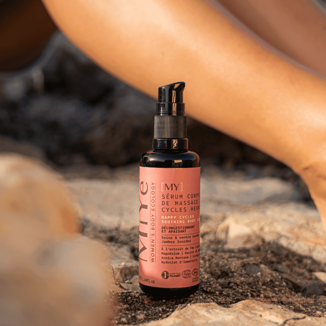 Le sérum corps posé sur des rochers, derrière lui on voit les jambes d'une femme qui se prélasse sur la plage.